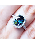 An oval-cut Australian sapphire ring for women, featuring a sparkling bluish-green hue, surrounded by a halo of diamonds set in 18K white gold, worn on a finger.