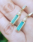 A closer look of a pair of 14k yellow gold emerald earrings placed on fingers against a blurred background.