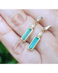 A closer look of a pair of 14k yellow gold emerald earrings placed on fingers against a blurred background.