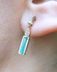 Close-up of a 14K gold baggette cut emerald stud earring worn on an ear.