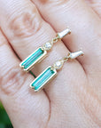 A 14k yellow gold emerald and diamond baggette cut stud earrings on fingers against a blurred green background.
