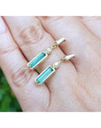 A 14k yellow gold emerald and diamond baggette cut stud earrings on fingers against a blurred green background.