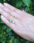 Hand displaying two gold emerald and diamond baggette cut stud earrings against a green leafy background