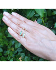 Hand displaying two gold emerald and diamond baggette cut stud earrings against a green leafy background