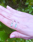 Hand displaying two emerald earrings with diamond and white gold against a green leafy background