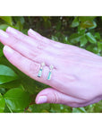 Hand displaying two emerald earrings with diamond and white gold against a green leafy background