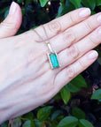 Hand displaying a gold pendant necklace with a natural emerald against a leafy background.