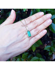 Hand displaying a gold pendant necklace with a natural emerald against a leafy background.