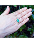 Baguette cut emerald pendant in 18K yellow gold with diamond accents, worn on a hand.
