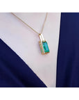 Baguette cut emerald pendant in 18K yellow gold with diamond accents, worn on a dark blue top.