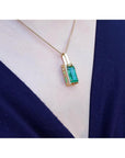 Baguette cut emerald pendant in 18K yellow gold with diamond accents, worn on a dark blue top.