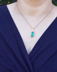 A woman wearing a navy blue outfit with a gold necklace featuring a baguette cut emerald pendant.