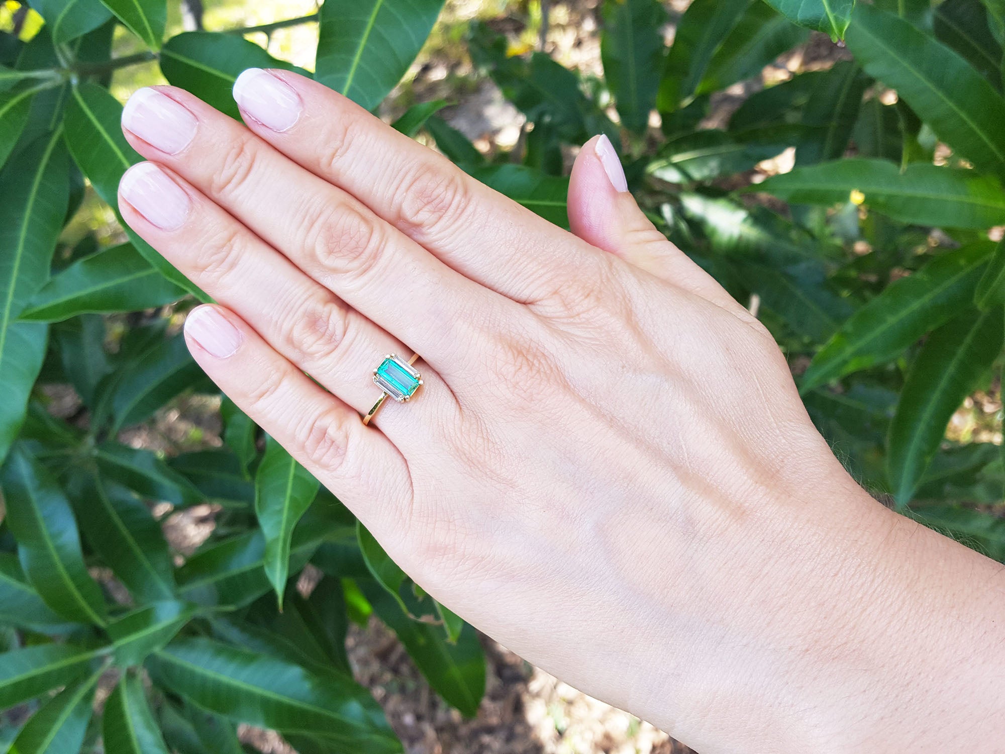 Hand wearing a baguette emerald and side diamonds ring with green leaves in the background.