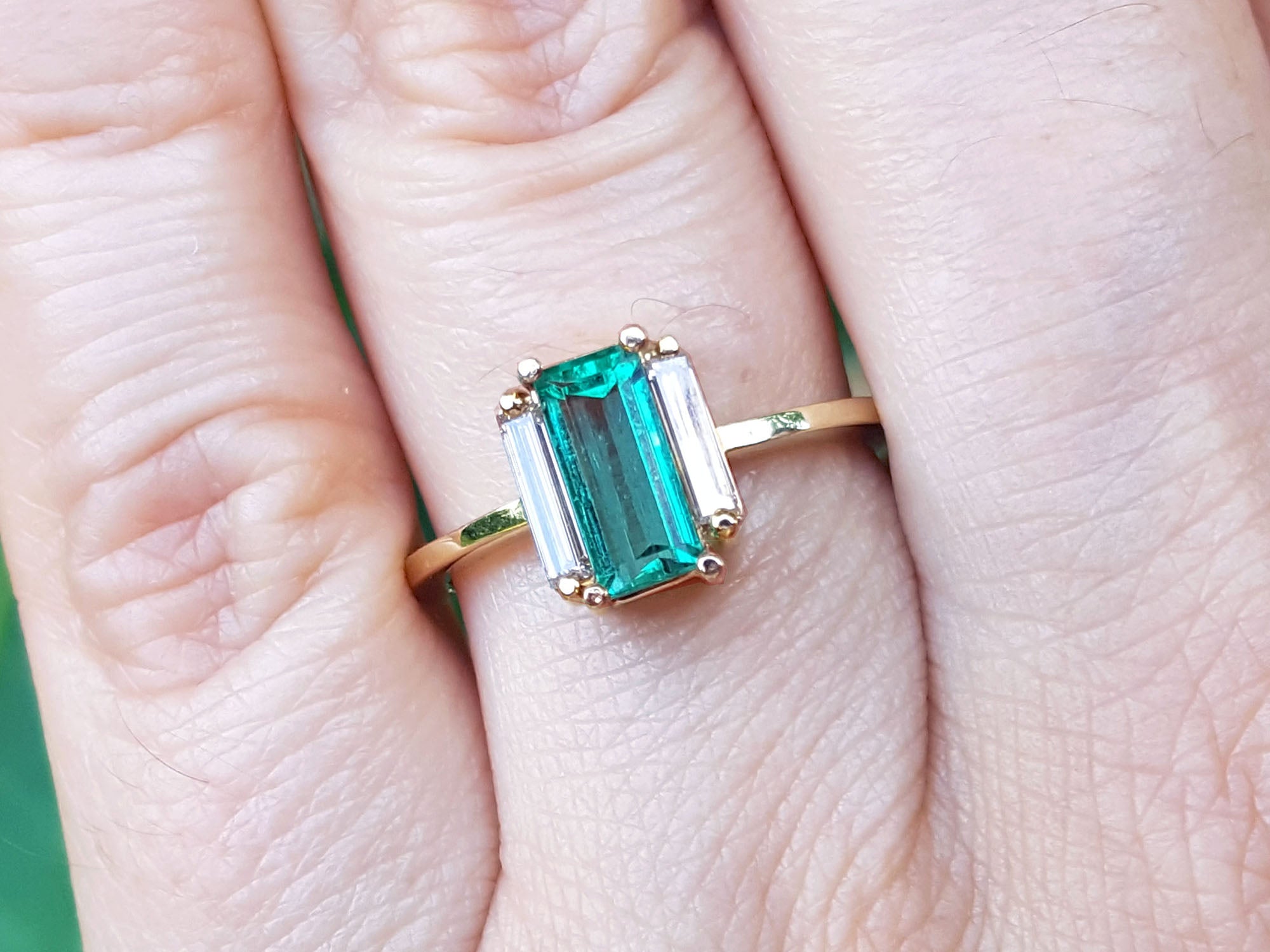 A baguette cut emerald ring with side diamonds on a finger with a soft background.