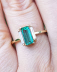 A baguette cut emerald ring with side diamonds on a finger with a soft background.