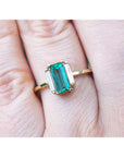 A baguette cut emerald ring with side diamonds crafted in 18k yellow gold on a finger.