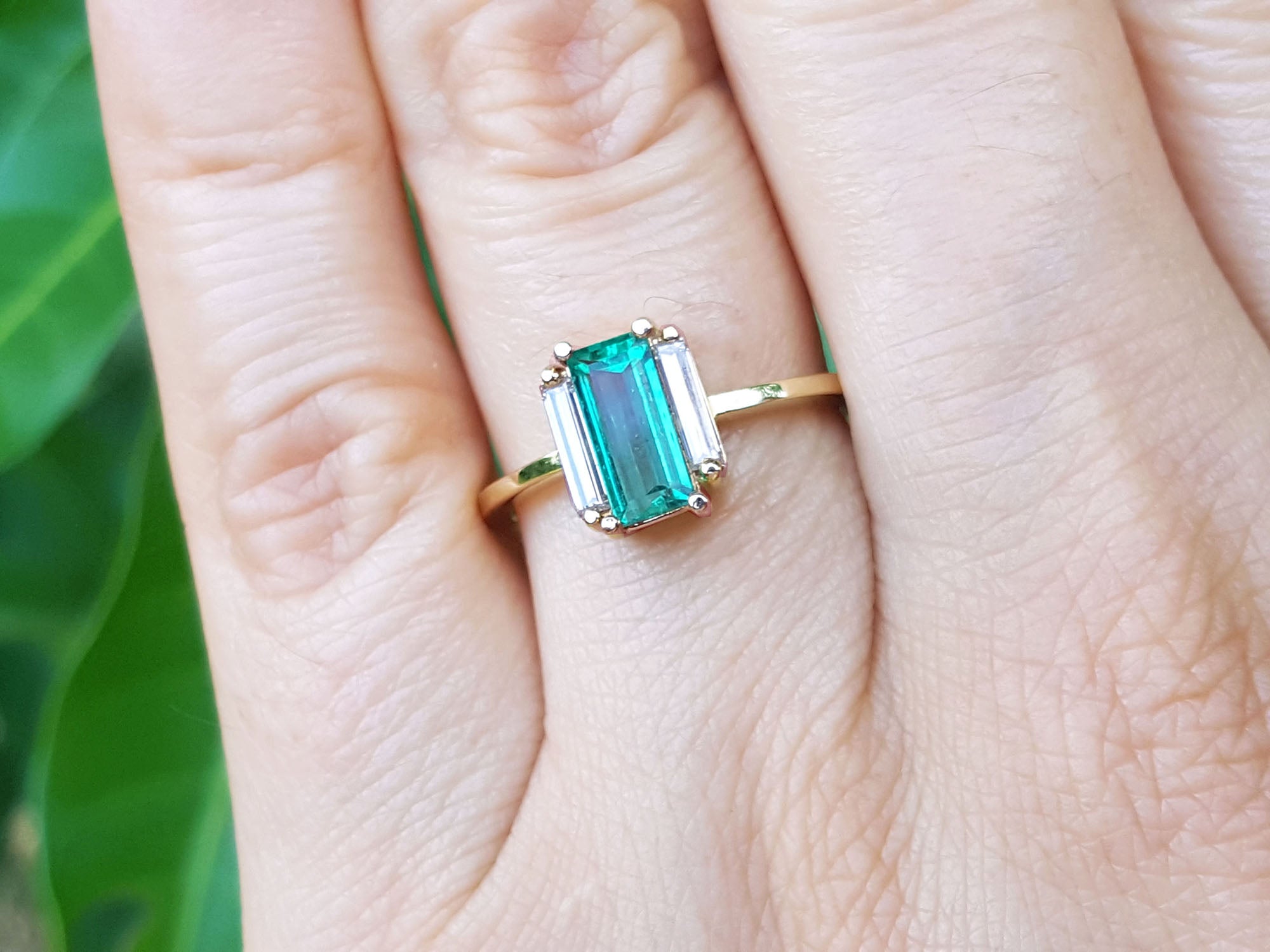Emerald and side straight baguettes in 18k gold ring on a person's finger against a blurred natural background.