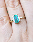 Emerald and side straight baguettes in 18k gold ring on a person's finger against a blurred natural background.