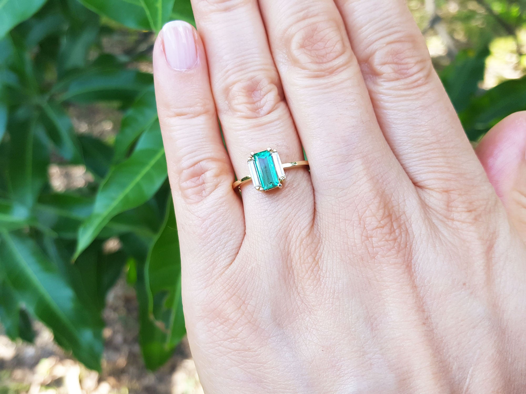 Hand wearing a ring with emerald baguette cut and diamonds each side against a leafy background.