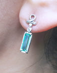 Close-up of a emerald baguette cut earring and diamond accent worn on an ear.
