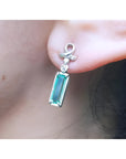 Emerald-cut natural emerald earrings in 14k white gold with ribbon design and accent diamonds.