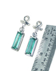 Pair of emerald baguette cut emerald earrings and white gold top ribbons next to a ruler for scale on a white background