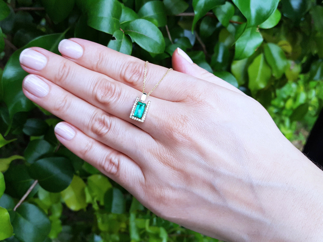 Hand displaying a gold pendant necklace with a natural emerald against a green leafy background.