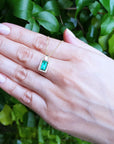 Hand displaying a gold pendant necklace with a natural emerald against a green leafy background.