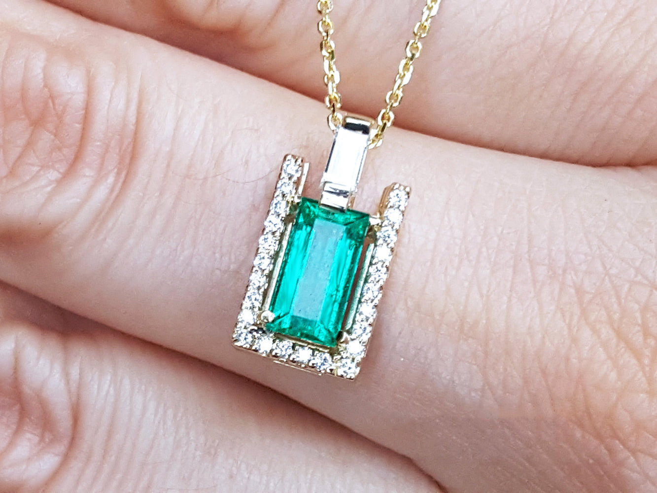 Emerald baguette cut gemstone pendant on a gold chain held between fingers.