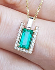 Emerald baguette cut gemstone pendant on a gold chain held between fingers.
