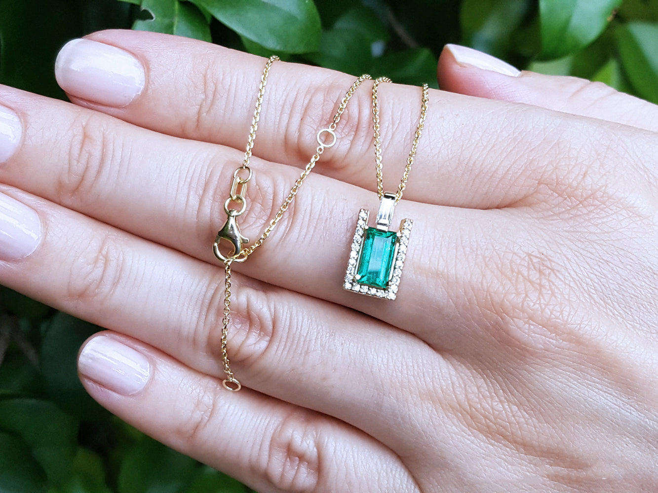 Gold necklace with a baguette cut emerald pendant held by a hand against a blurred green leafy background.