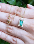 Gold necklace with a baguette cut emerald pendant held by a hand against a blurred green leafy background.