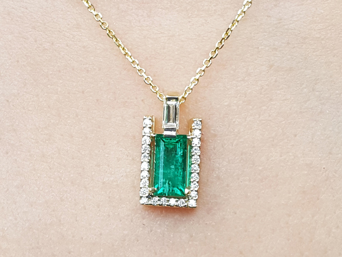 Emerald baguette pendant with diamond accents on a gold chain against a beige skin background.