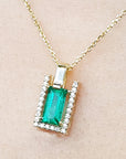 Gold necklace with a baguette cut emerald pendant on a beige skin background.