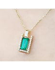 Gold necklace with a baguette cut emerald pendant on a beige skin background.