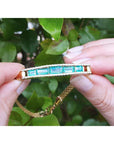 14K gold bracelet with five vibrant bluish-green emeralds and surrounding diamonds, held against green leaves.