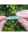 14K gold bracelet with five vibrant bluish-green emeralds and surrounding diamonds, held against green leaves.