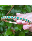Gold bracelet with round cut emeralds held by a hand between fingers against a green leafy background.