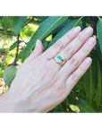 Hand wearing a gold ring with a natural colombian emerald against a green leafy background.