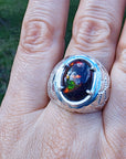 Men's silver ring with a colorful black opal held between fingers against a blurred green background