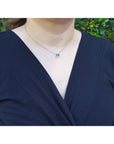A lady wearing a navy blue top with a necklace featuring a blue sapphire gemstone against a blurred green background