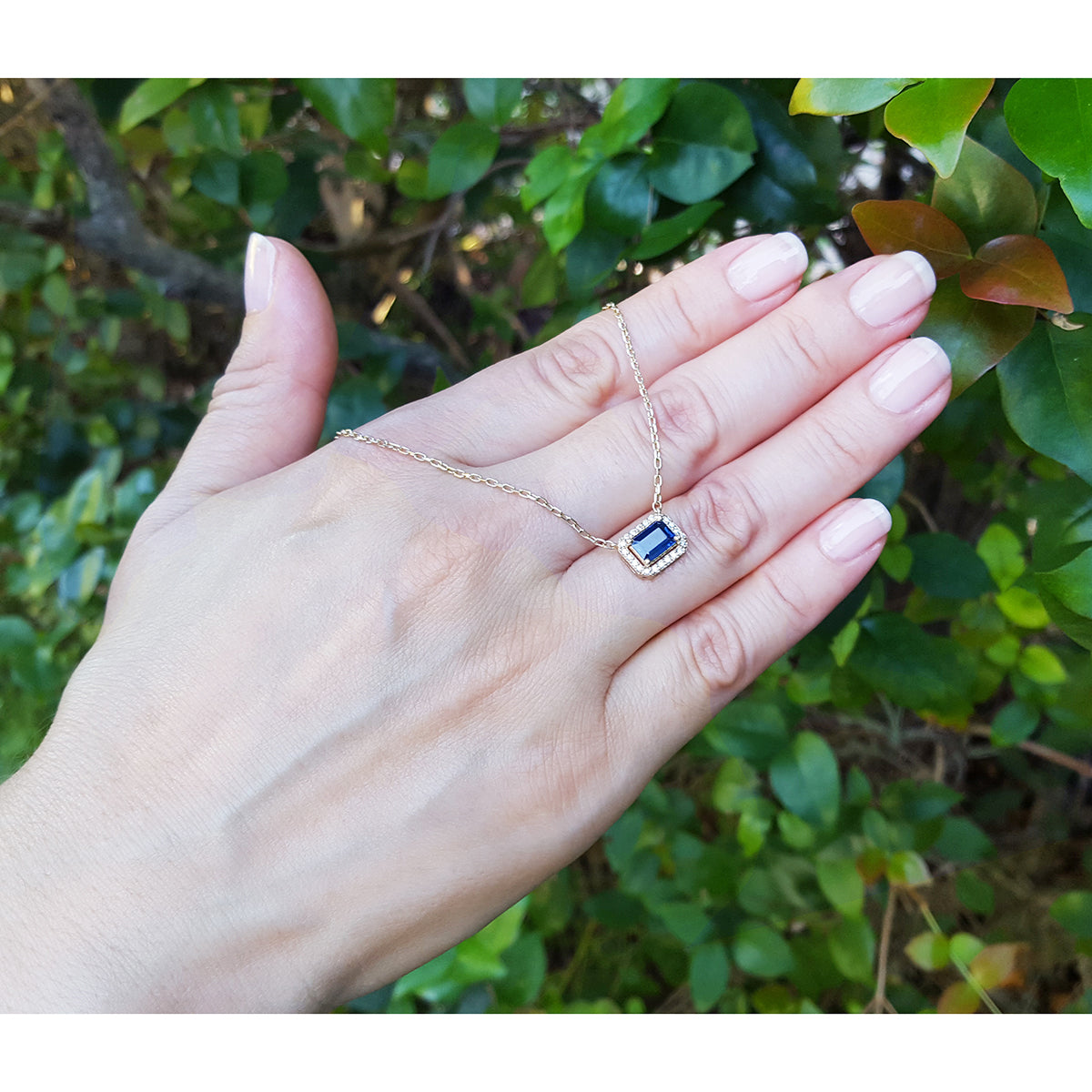 Hand showcasing a gold necklace with a blue sapphire gemstone against a green leafy background