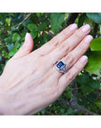 Women's 18K gold ring with a 4.51 ct emerald cut blue sapphire and three-row diamond band on a hand.