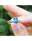 Gold ring with a blue sapphire held between fingers against a green leafy background