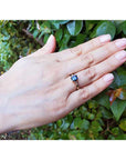 Hand wearing a gold ring with a blue sapphire against a green leafy background