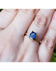 A 14k yellow gold ring with a blue natural sapphire rich medium-dark hue, flanked by two tapered baguette natural diamonds on a person's finger.