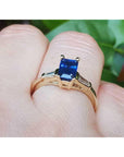 Yellow gold ring with a blue sapphire on a finger against a blurred green background