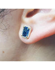 Emerald cut blue sapphire stud earring with halo diamonds in 14K yellow gold on an ear.