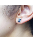 Emerald cut blue sapphire halo diamond stud earring in 14K yellow gold on an ear.