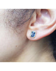 Emerald cut blue sapphire halo diamond stud earring in 14K yellow gold on an ear.